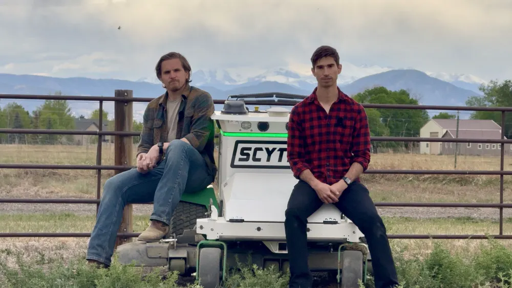 Jack Morrison and Davis Foster sitting on a Scythe autonomous mower in a Colorado field, the Front Range behind them